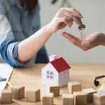 Gros plan d'une main de femme qui donne la clé d'une maison à un homme au dessus d'une tzable en bois sur laquelle est posée une maison miniature en bois