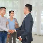 Un promoteur immobilier en costume serre la main d’un couple souriant dans un intérieur lumineux.