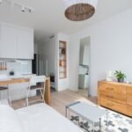 Un studio de style scandinave avec une cuisine blanche intégrée, une table haute en bois servant de bar, un canapé blanc et une commode en bois clair