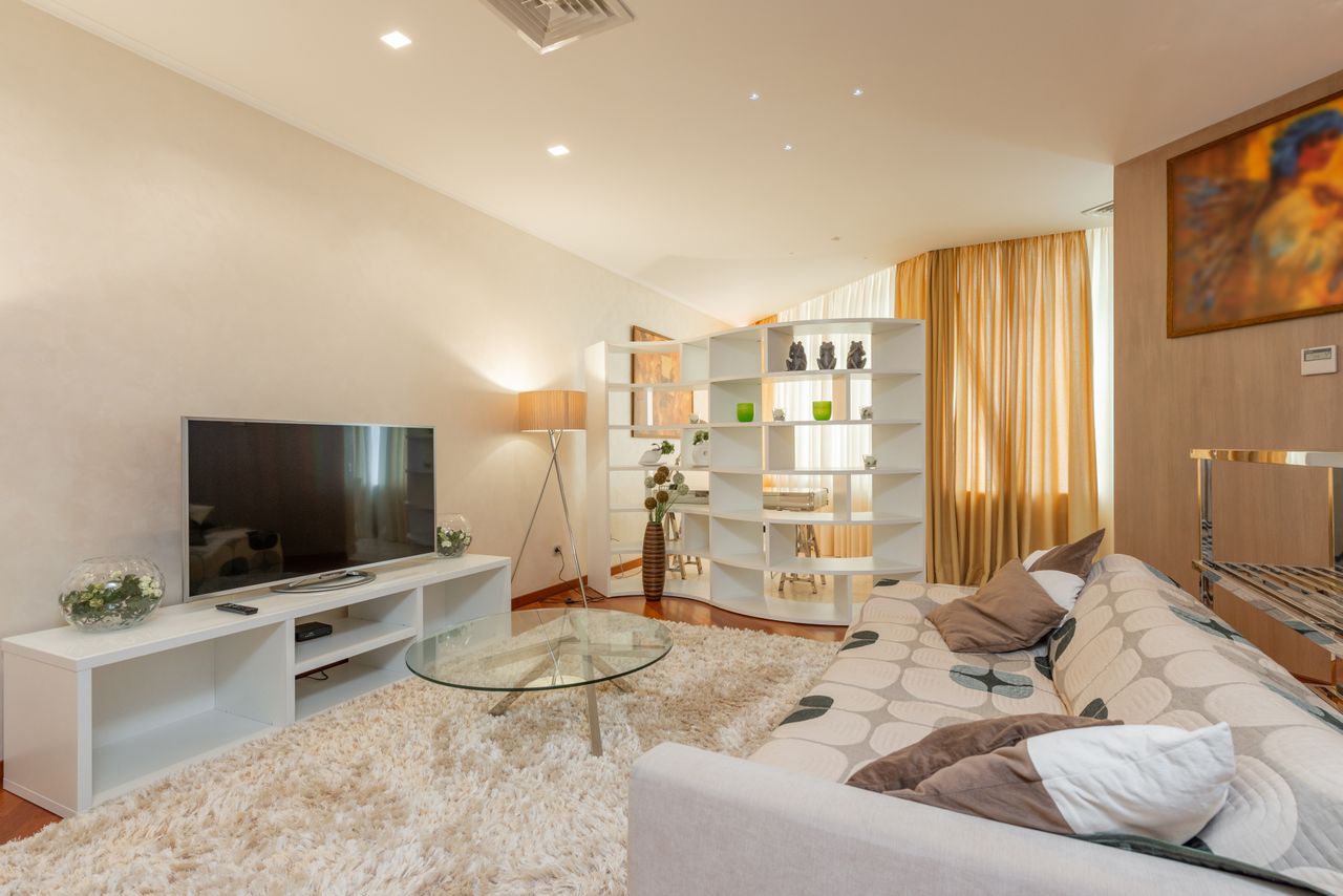 Cosy Home un spécialiste de la location meublée au service des propriétaires et des entreprises un grand salon lumineux comprenant un canapé aux motifs géométriques, une table basse en verre, un meuble TV blanc avec un écran plat, un tapis moelleux et une étagère séparatrice